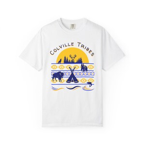 Könnte beinhalten: Ein weißes Kurzarm-T-Shirt mit einer Grafik und dem Text Colville Tribes. Das Design zeigt eine gelbe Sonne, eine Waldsilhouette, einen Hirsch, einen Bären, einen Bison, ein Tipi und einen Fisch, akzentuiert durch blaue und gelbe geometrische Muster.