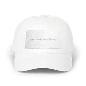 Puede incluir: Gorra de béisbol blanca con visera curva. La gorra presenta un parche rectangular con el texto "Good Food > Good Photos" en gris claro. La gorra está hecha de una tela lisa y tiene un botón en la parte superior.