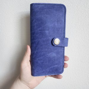 May include: A rectangular blue leather wallet featuring a distressed texture, visible dark stitching along the edges, and a small tab closure secured with a circular metallic snap button embossed with a paw print and text.
