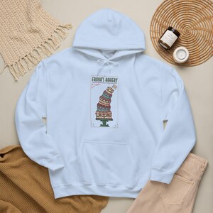 Puede incluir: Sudadera con capucha azul claro con un gráfico de una tarta de varios pisos sobre un soporte, con el texto "Fauna's Bakery". El pastel tiene varios colores y patrones. La sudadera con capucha tiene una capucha con cordón y un bolsillo delantero.