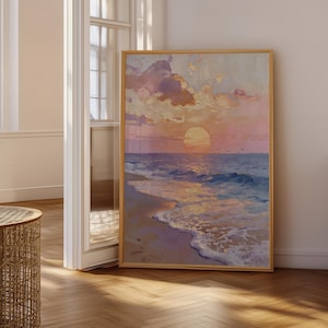 May include: A framed painting depicting a beach scene at sunset. The artwork shows a colourful sky with pink, orange, and yellow hues, mirrored in the ocean waves. The painting is displayed in a room with natural light and wooden flooring.