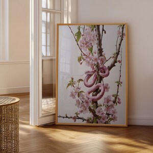 Könnte beinhalten: Ein rechteckiger gerahmter Wandkunstdruck mit einer detaillierten Illustration einer rosa Schlange, die sich um einen blühenden Baumzweig mit hellrosa Blüten und grünen Blättern vor einem schlichten, hellen Hintergrund windet.