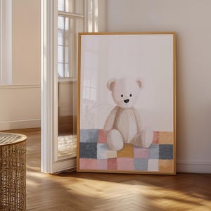 Puede incluir: Una lámina de arte mural enmarcada que muestra un oso de peluche de color beige claro sentado sobre una colcha de retazos colorida. La colcha está compuesta por parches cuadrados en tonos azul, amarillo, rosa y crema. El oso tiene nariz negra.