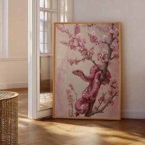 Pode incluir: Uma impressão de arte de parede vertical emoldurada apresentando uma cobra rosa enrolada em torno de um galho de cerejeira em flor. A obra de arte exibe uma paleta de cores rosa monocromática suave com texturas detalhadas nas escamas da cobra.