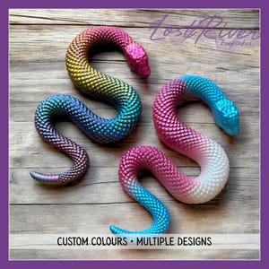 May include: Two articulated snake figures with textured scales. The top snake features a gradient of yellow, orange, and blue. The bottom snake displays a gradient of pink, white, and blue. Text reads Lost River Keepsakes and CUSTOM COLOURS.