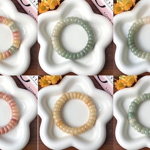 Puede incluir: Seis pulseras de cuentas coloridas en tonos rosa, verde, amarillo y blanco se exhiben en platos blancos con forma de flor. Las pulseras tienen una textura acanalada y están dispuestas en forma circular.