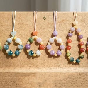 Peut inclure: Six ornements perlés colorés sont suspendus à un présentoir en bois. Chaque ornement présente une forme de goutte d'eau composée de petites perles rondes de différentes couleurs, dont le vert, le bleu, le violet et l'orange. Les ornements sont enfilés sur de fins cordons.