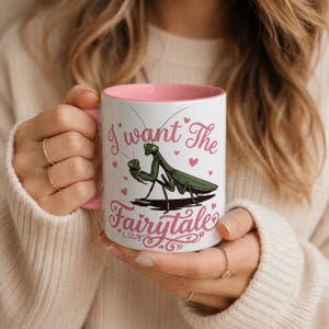 May include: A white ceramic mug with a pink interior and handle, featuring a green praying mantis illustration and the pink text "I want The Fairytale" surrounded by small pink hearts on the front surface of the mug.
