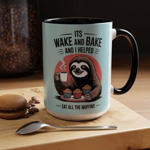 Könnte beinhalten: Keramik-Kaffeetasse mit schwarzem Henkel und Rand, mit einer Illustration eines Faultiers, das eine Kaffeetasse und Muffins hält. Die Tasse trägt den Text "ITS WAKE AND BAKE AND I HELPED - EAT ALL THE MUFFINS" auf hellblauem Hintergrund.
