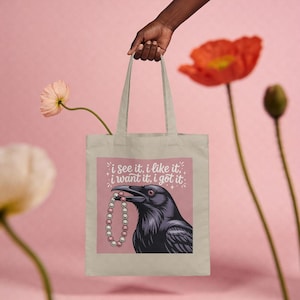 Puede incluir: Bolso tote beige con un cuervo sosteniendo un collar de perlas, con el texto "i see it, i like it, i want it, i got it" en un cuadrado rosa. El bolso es sostenido por una mano, con flores en el fondo.
