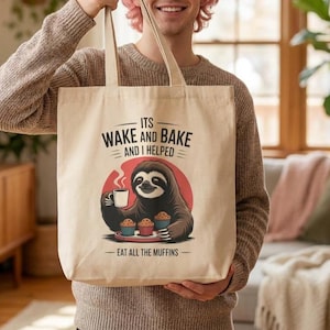 Könnte beinhalten: Beige Canvas-Tragetasche mit einer Illustration eines Faultiers, das eine Kaffeetasse und Muffins hält. Der Text auf der Tasche lautet "Its Wake and Bake and I Helped - Eat All The Muffins". Die Tasche hat ein schlichtes Design mit hellem Hintergrund.