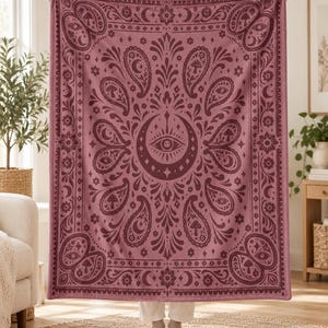 May include: A rectangular throw blanket in shades of dusty rose and deep maroon, featuring a central crescent moon and eye motif surrounded by symmetrical paisley patterns, floral elements, and small mushroom illustrations.