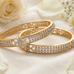 May include: Two gold bangles featuring a central flower-shaped motif and rows of small clear stones. The circular bracelets have a polished metallic finish and intricate cut-out detailing along the edges, creating a structured and ornate appearance.