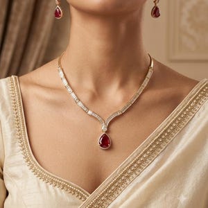 Op de afbeelding: Een gouden ketting met een centrale peervormige rode edelsteen hanger omringd door kleine witte steentjes, met een ketting van witte stenen in baguette-slijpvorm. Bijpassende peervormige rode oorbellen met witte steentjes als accent.