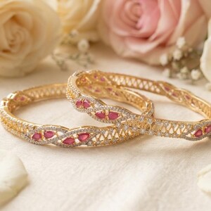May include: Two gold bangles featuring an intricate twisted metal design, embellished with small clear stones and teardrop-shaped red gemstones. The bracelets have a hinged construction and a polished finish, showcasing a detailed lattice pattern.