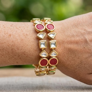 Op de afbeelding: Twee goudkleurige armbanden met een herhalend patroon van ronde rode stenen afgewisseld met heldere, geometrisch gevormde stenen. De armbanden hebben een gepolijste metalen afwerking en zijn ontworpen met een reeks verbonden, hoekige zettingen.