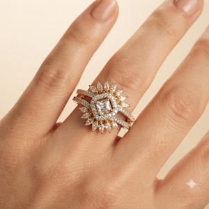 May include: A gold ring set featuring a central princess-cut stone surrounded by a halo of small round stones. The band is accented with pavé stones and a fan-shaped arrangement of marquise-cut stones extending from the center setting.
