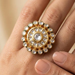 May include: A gold-toned ring featuring a large, circular centerpiece with a central white stone surrounded by a ring of smaller white stones, all encased in a scalloped gold frame with an outer border of additional white stones.