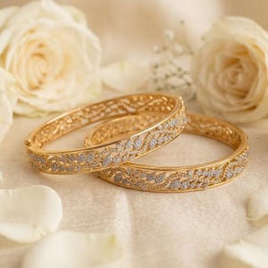 May include: Two gold-toned hinged bangle bracelets featuring an intricate filigree pattern with embedded clear stones. The bracelets have a polished metallic finish and a curved, circular shape, showcasing a detailed decorative design throughout.