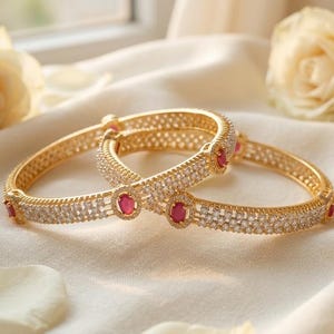 Puede incluir: Dos brazaletes dorados con una fila central de piedras rojas ovaladas rodeadas por pequeños cristales transparentes. Las bandas de metal tienen un patrón texturizado similar a una rejilla en los bordes, creando una apariencia detallada y ornamentada.