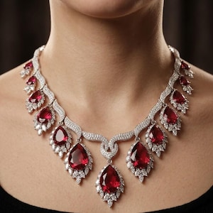 Puede incluir: Un collar deslumbrante con grandes piedras preciosas rojas en forma de lágrima, cada una rodeada de piedras blancas más pequeñas y brillantes. El collar está engastado en un metal plateado y descansa elegantemente sobre el cuello de una persona, contra una blusa negra.