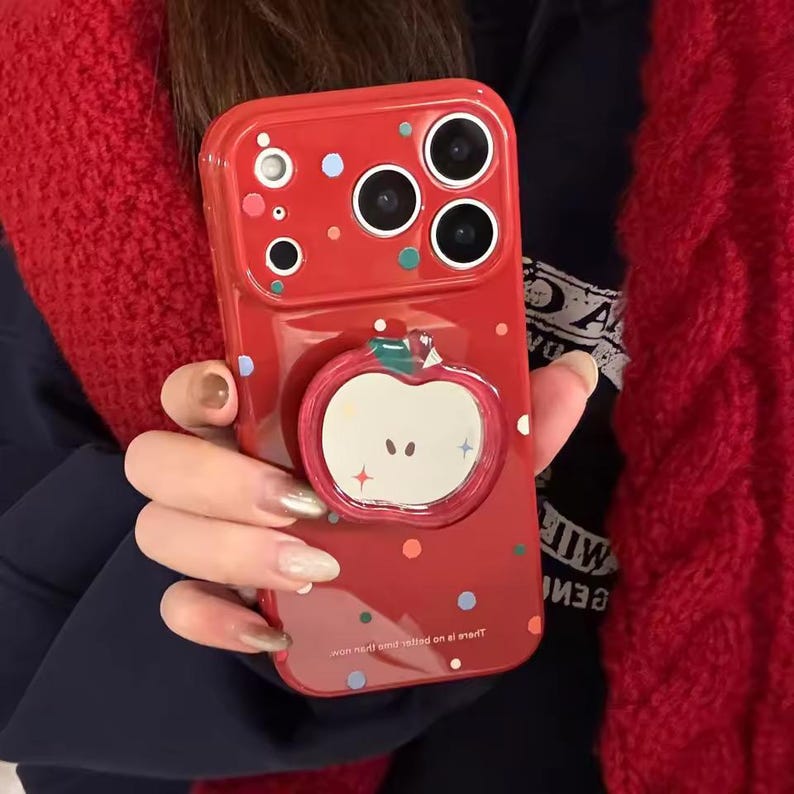 May include: A red phone case with an apple-shaped grip. The case has a glossy finish and features colorful polka dots. The apple grip is white with a red outline and a green stem. The case has a camera module with three lenses.