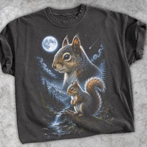 May include: A charcoal grey short-sleeve t-shirt featuring a graphic print of two squirrels in a forest landscape at night. The scene includes a full moon, a starry sky, a waterfall, and pine trees, rendered in shades of brown, white, and blue.