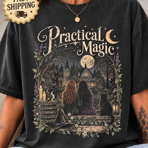May include: A charcoal gray t-shirt featuring a graphic print with the text Practical Magic. The design shows two figures sitting before a house under a full moon, with a stack of books labeled Spells, Herbs, Sisters, Always, a cat, and a witch hat.