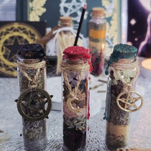 May include: Three glass bottles filled with herbs and crystals, each sealed with a different colored wax. The bottles are adorned with twine and metal charms, including a pentacle and a crescent moon. The background includes a book titled "Witch's Way".