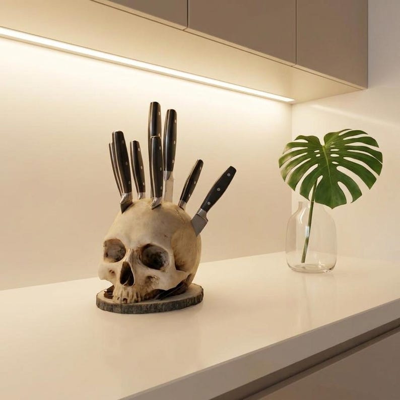 May include: A novelty knife block shaped like a human skull, holding several black-handled knives. The skull is off-white and rests on a wooden base. A decorative Monstera leaf in a glass vase is in the background.