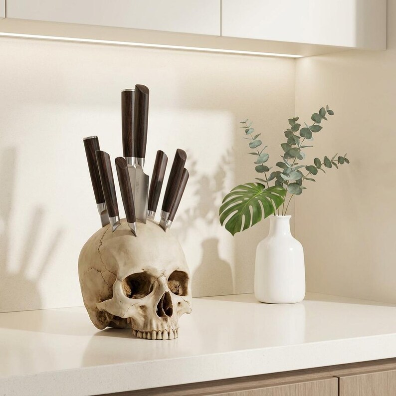 May include: A decorative skull-shaped knife holder with several knives inserted into the top. The knives have dark brown handles and silver blades. The skull is off-white and sits on a white countertop. A white vase with greenery is to the right.