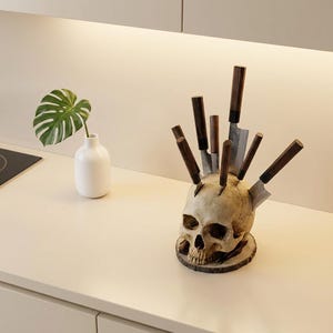 May include: A decorative skull-shaped knife holder with several knives inserted into it. The skull is off-white with dark eye sockets and a detailed jawline. The knives have dark wooden handles and silver blades. The holder sits on a wooden base.