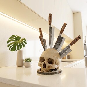 May include: A skull-shaped knife holder with six knives inserted. The knives have wooden handles and silver blades. The skull is off-white and sits on a wooden base. The kitchen setting includes a monstera leaf in a vase.
