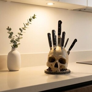 May include: A kitchen knife set displayed in a skull-shaped knife block. The skull is off-white with dark eye sockets and a dark wooden base. The knives have black handles and silver blades. A white vase with greenery is to the left.