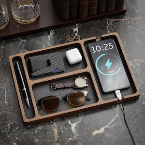 May include: A wooden desk organizer with multiple compartments. It holds a charging smartphone, wallet, watch, sunglasses, pen, and wireless earbuds. The phone displays 10:25 and 65% battery. The organizer sits on a marble surface.