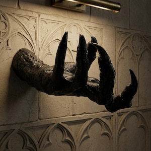 Könnte beinhalten: Eine dunkle, strukturierte Krallenhand-Skulptur, die aus einer hellen Wand hervortritt. Die Hand hat lange, scharfe Krallen und ist so positioniert, als würde sie sich ausstrecken. Die Wand hat ein dekoratives Muster und einen Riss.