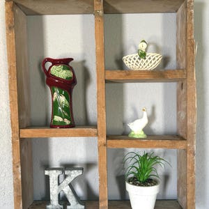 May include: A rustic wooden shelf unit with four compartments, each holding decorative items. Includes a red and green vase, a white ceramic swan in a basket, a white ceramic goose, a metal letter "K", and a potted plant.