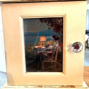Peut inclure: Un petit meuble blanc cassé avec une image encadrée d'une femme assise à une table au bord d'un lac. Le meuble a un bouton en cristal et une peinture écaillée.