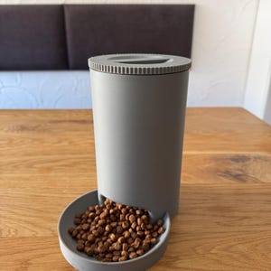 Puede incluir: Una taza de cerámica blanca con un asa negra y el texto "But First, Coffee" en un estilo manuscrito. La taza está sobre una superficie blanca. La taza mide aproximadamente 10 cm de alto.