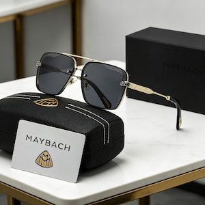 May include: Square sunglasses with a gold metal bridge and temples, black rims, and dark grey lenses. The sunglasses sit on a black leather case with a gold emblem and a white card with the brand name "MAYBACH".