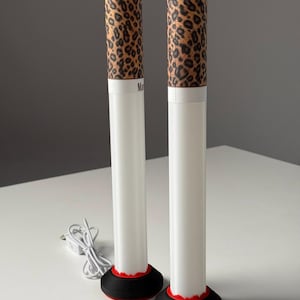 May include: Two white cylindrical lamps with a leopard print top and a black base with a red accent. The lamps are plugged in with a white cord. The text "Ma" is visible on the lamps.