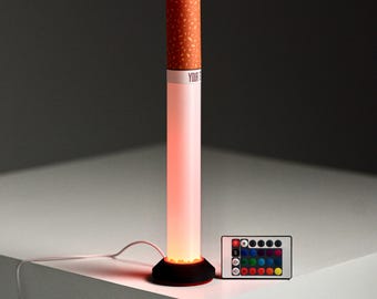 May include: A cylindrical LED lamp shaped like a cigarette with a speckled orange filter top and a white body that glows with a soft red light. The lamp sits on a black circular base with a power cord and includes a small remote control with buttons.