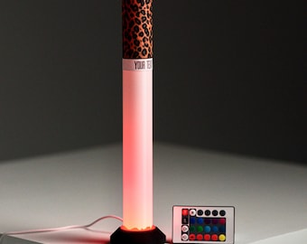 May include: A cylindrical LED lamp with a leopard print top section and a white band featuring the text YOUR TEXT. The lamp emits a red glow from its base, which is black and circular. A small rectangular remote control with colored buttons is included.