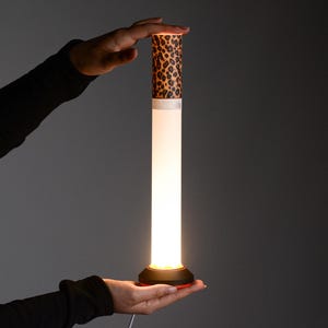 Op de afbeelding: Een hoge, cilindervormige LED-lamp met een witte, verlichte centrale buis. Het bovenste gedeelte heeft een bruin met zwart luipaardpatroon. De voet is zwart met een dunne rode accentring aan de onderkant en verbonden met een stroomkabel.