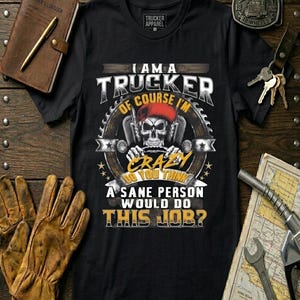Puede incluir: Camiseta negra con el texto "I am a trucker, of course I'm crazy. Do you think a sane person would do this job?" Una calavera con un sombrero rojo está en el centro del diseño. Otros artículos incluyen un diario de cuero, guantes y una hebilla.
