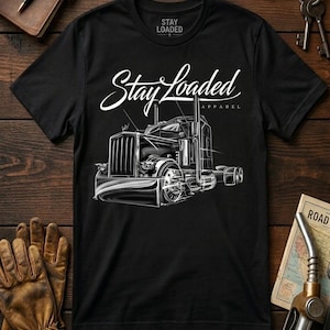 Puede incluir: Camiseta negra con un gráfico blanco detallado de un camión. La camiseta presenta el texto "Stay Loaded Apparel" en una fuente estilizada. La imagen está sobre un fondo de madera con guantes de cuero, un diario y llaves.