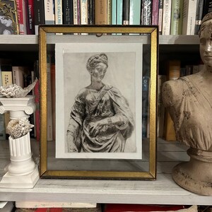 Pode incluir: Uma impressão emoldurada em preto e branco de um busto clássico, em um fundo de livros. A moldura é dourada e a impressão retrata uma mulher em trajes clássicos. A imagem é exibida em uma prateleira com vários livros.