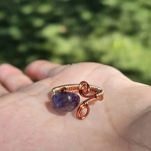 May include: A wire-wrapped ring featuring a polished, deep purple amethyst stone. The band is constructed from coiled copper wire, with one end terminating in a decorative spiral loop and the other securing the gemstone in a simple setting.