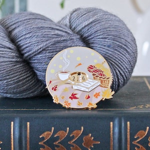 以下が含まれることがあります： コーヒーカップ、本、糸かごが描かれた秋のシーンが特徴の丸いエナメルピン。灰色の糸玉とヴィンテージ本の背景に配置されています。ピンは金色の縁取りで、秋の葉で飾られています。