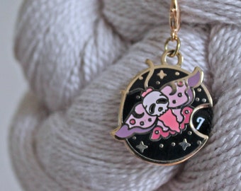 May include: A gold-colored enamel stitch marker featuring a moth design with a skull, pink wings, and a black background with stars. The marker is attached to a gold-colored clasp and is set against a backdrop of gray yarn.
